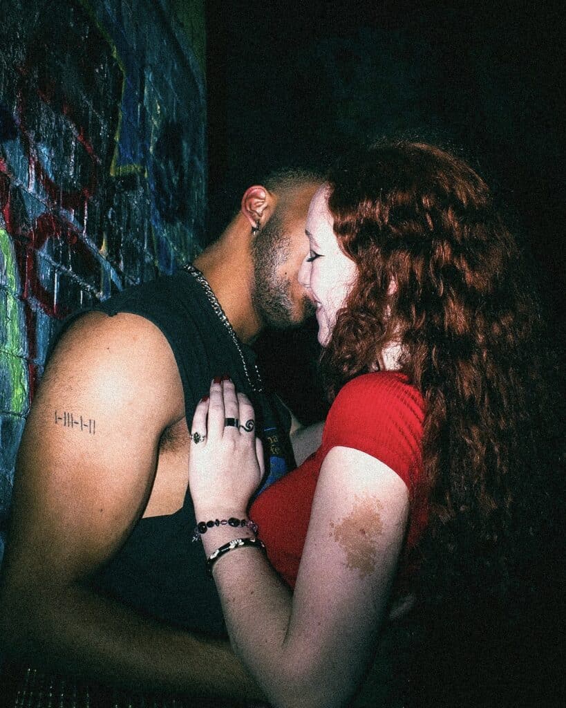 A man and woman stand close together against a graffiti-covered wall at night, appearing intimate. The man wears a sleeveless shirt, and the woman with curly red hair smiles gently, her hand on his chest.