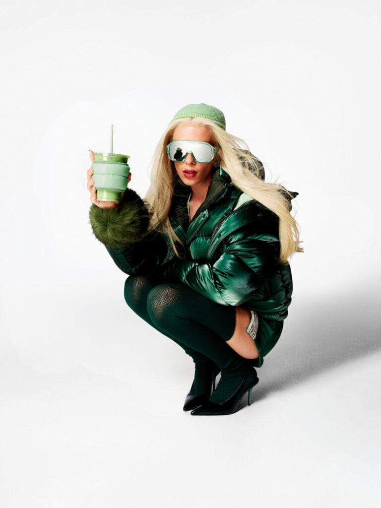 A person with long blonde hair squats on the ground wearing a shiny green puffer jacket, dark tights, black heels, large reflective sunglasses, and a green beanie, holding a green cup with a straw against a white background.