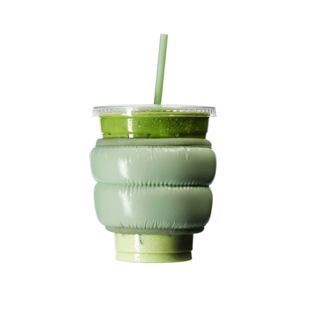 A green smoothie in a clear plastic cup with a green straw is wrapped in a puffy, light green insulated sleeve, set against a white background.