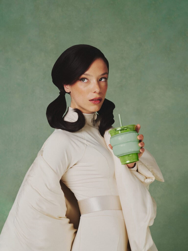 A woman with dark, styled hair and a serious expression wears a light, futuristic outfit and holds a green collapsible cup with a straw against a plain green background.