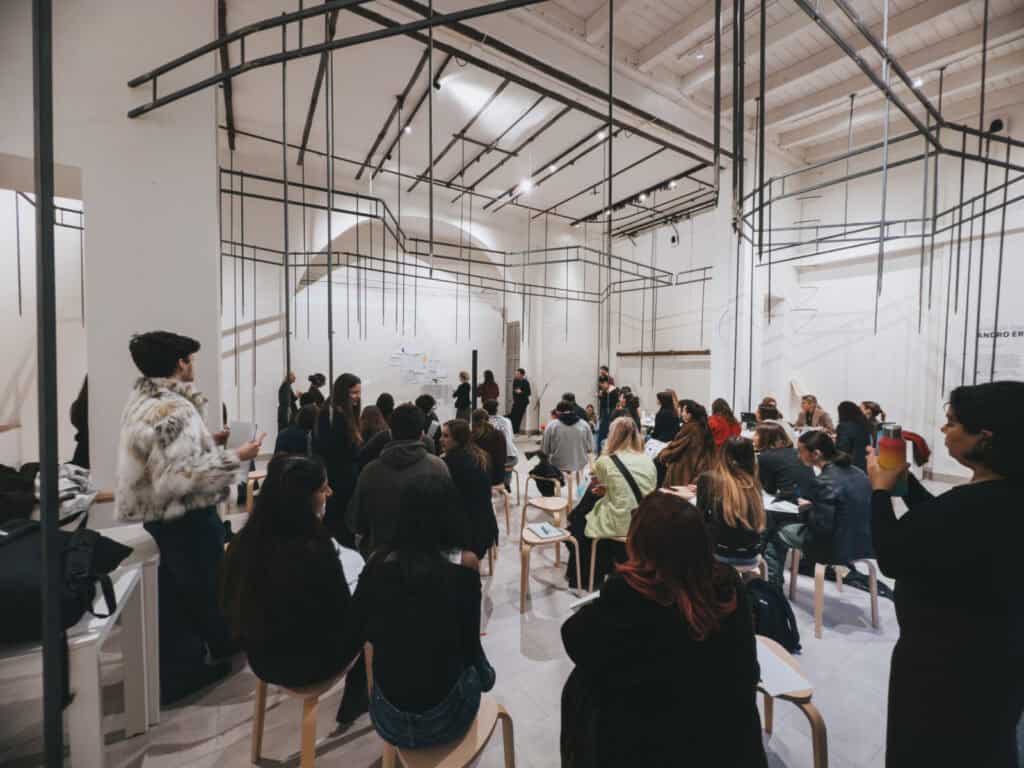 A group of people sits on stools in a bright, modern room, attentively listening to someone speaking at the front. Some are taking notes, while others face forward. The ceiling features abstract black rod installations.