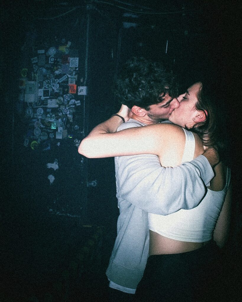 A young couple passionately kisses and embraces in a dimly lit room with a sticker-covered wall in the background. The mood is intimate and slightly moody, with shadowy lighting.