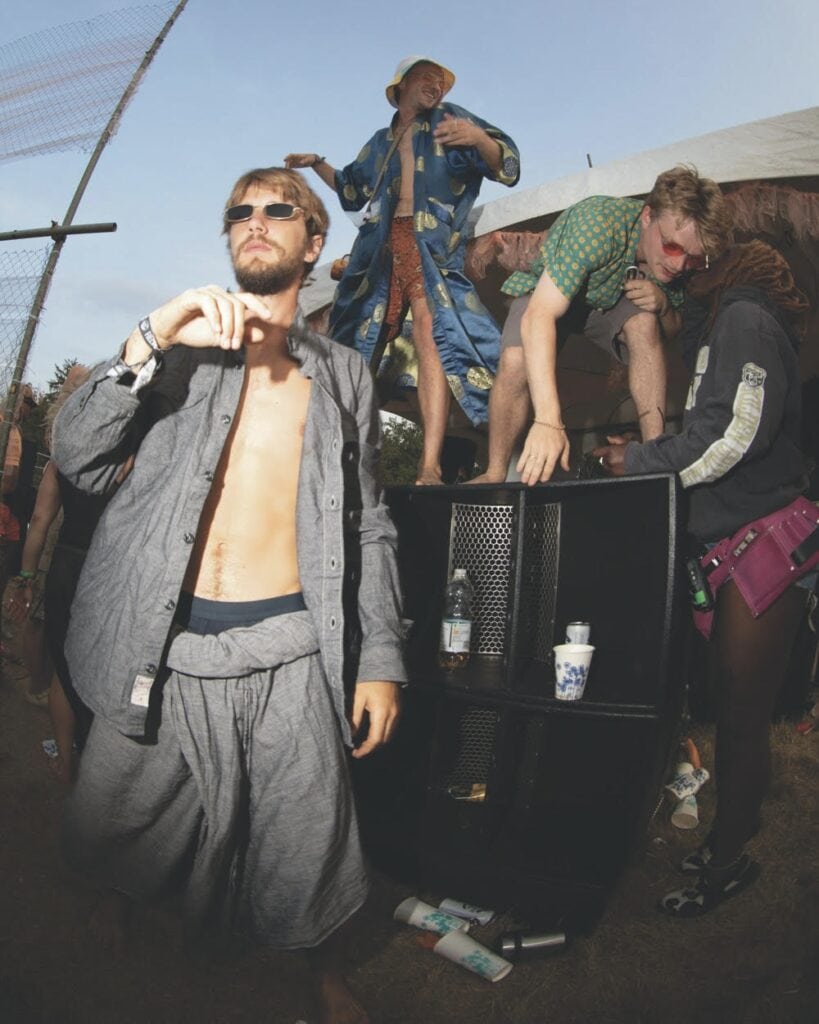 Three people at an outdoor event wear casual and eccentric clothing. One person stands confidently in the foreground, shirtless and wearing sunglasses, while two others interact with a black speaker in the background. Cups and bottles are on the ground.