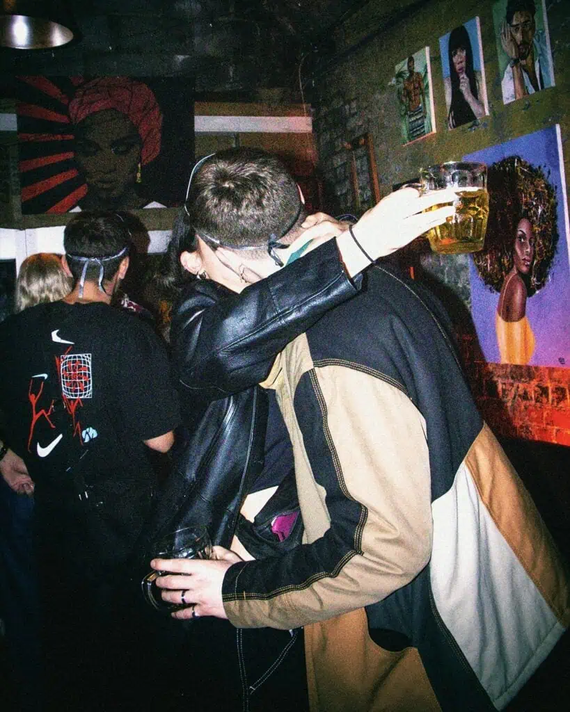 Two people hug in a dimly lit room with drinks in hand, surrounded by vibrant artwork on brick walls. Several other people are visible in the background, enjoying the lively, colorful atmosphere.