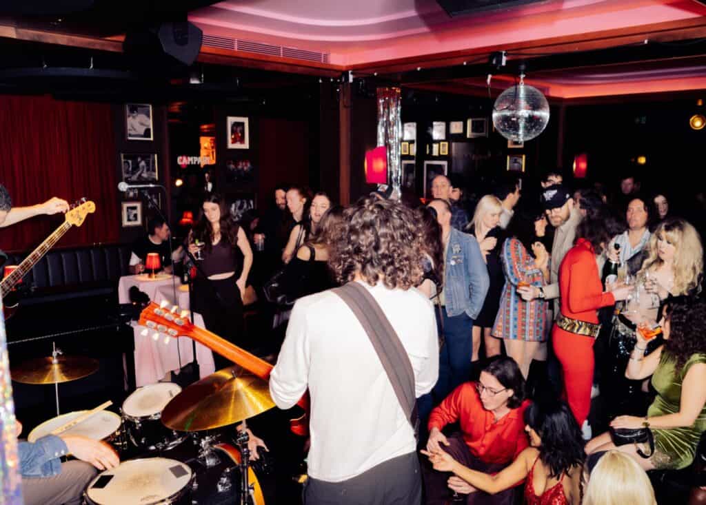 A band performs in a lively, crowded nightclub as people dance, talk, and watch. The room is decorated with red curtains, framed photos, and a disco ball hangs from the ceiling. The atmosphere is energetic and festive.
