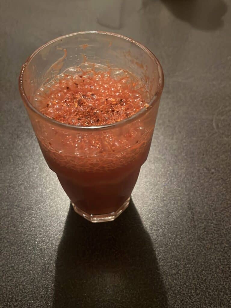 A glass filled with a reddish, frothy beverage is placed on a dark surface, with some black seasoning sprinkled on top.