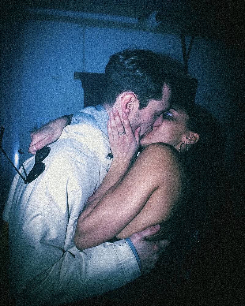A couple is passionately kissing in a dimly lit room. The man is wearing a light jacket and glasses, while the woman has long dark hair and is embracing him closely. The atmosphere is intimate and emotional.