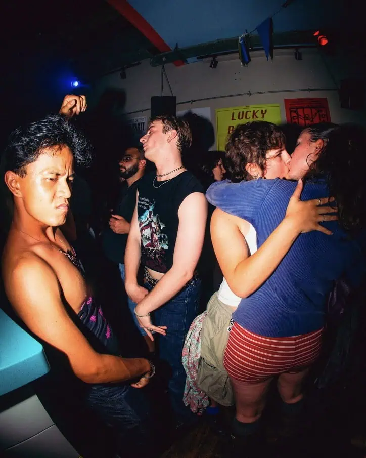 A group of people in a dimly lit club; one person dances energetically in the foreground, while two people kiss and others chat in the background. Colorful posters are visible on the wall.