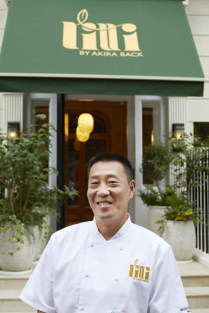 A smiling chef in a white uniform stands in front of a restaurant entrance with a green sign reading Fili by Akira Back. Two large potted plants flank the doorway.