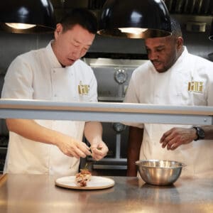 Two chefs in white uniforms prepare a dish together in a professional kitchen. One chef adds garnish to food on a plate, while the other watches, standing beside a metal mixing bowl.