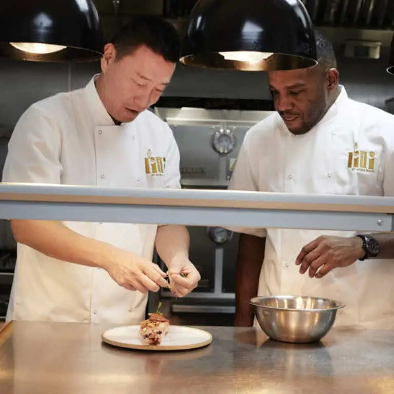 Two chefs in white uniforms prepare a dish together in a professional kitchen. One chef adds garnish to food on a plate, while the other watches, standing beside a metal mixing bowl.