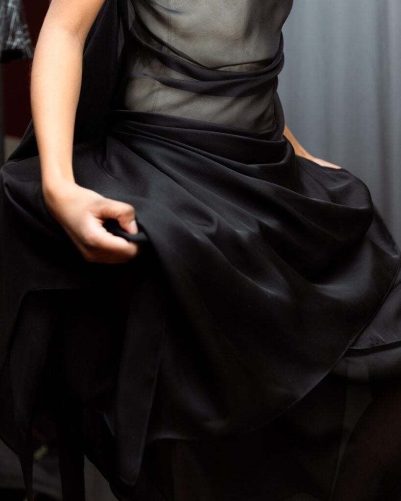 A person in a sheer black dress holds the flowing fabric of the skirt with one hand. The upper part of the dress is semi-transparent, and the background is out of focus.