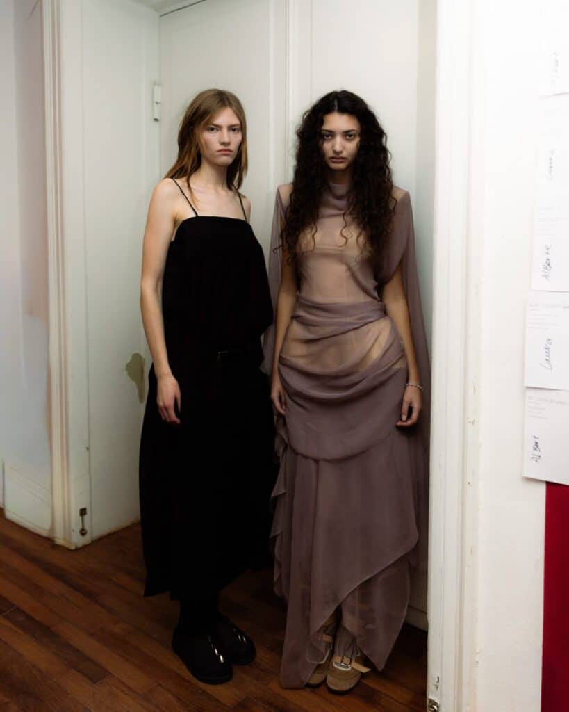 Two women stand side by side indoors. One wears a long black Anne Sofie Madsen dress with thin straps and boots, the other wears a sheer taupe gown with draped layers and beige shoes. Both have serious expressions and long hair.