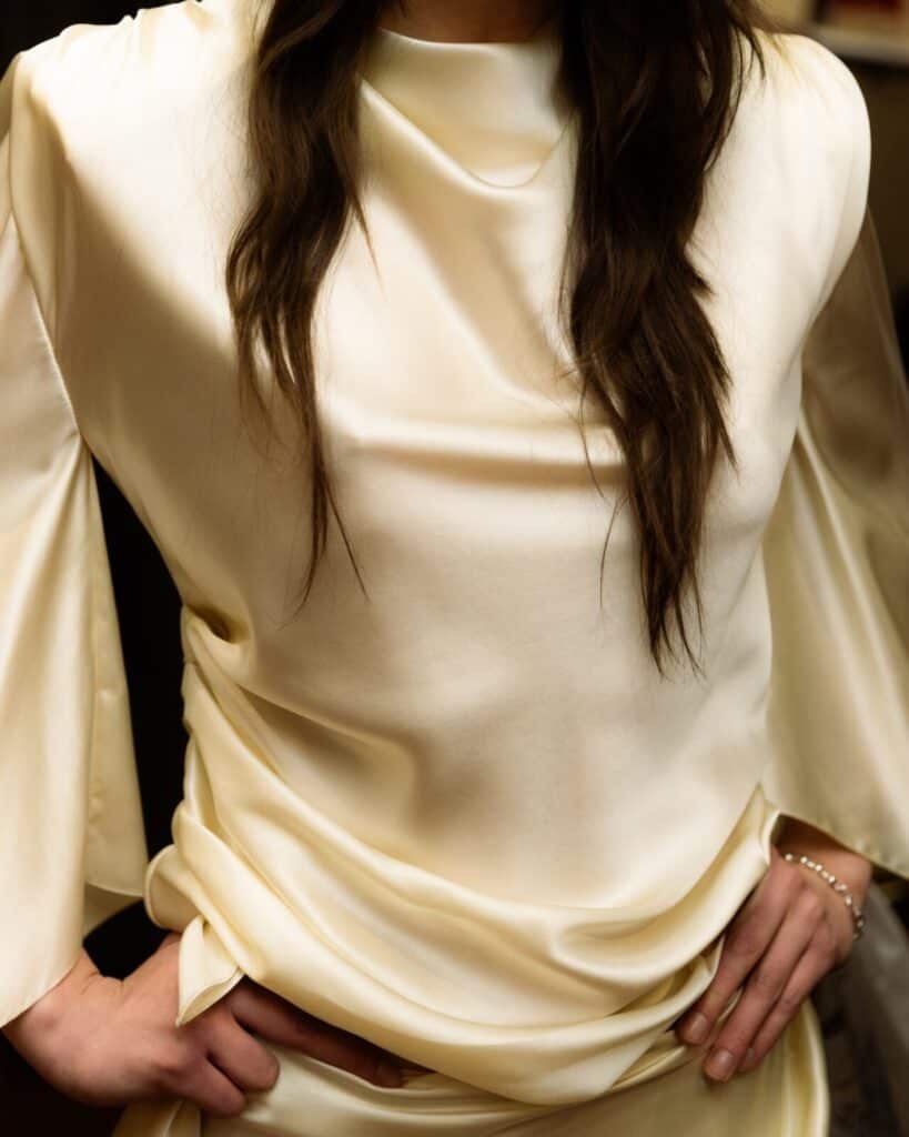 A person with long, wavy brown hair wears a shiny, cream-colored satin Anne Sofie Madsen blouse with draped fabric and wide sleeves, standing with hands on hips. Their face is not visible.