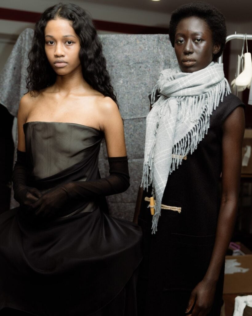 Two models stand indoors; one wears an Anne Sofie Madsen strapless black dress with sheer black gloves, and the other wears a black sleeveless outfit with a light gray scarf. They pose in front of a textured fabric backdrop.