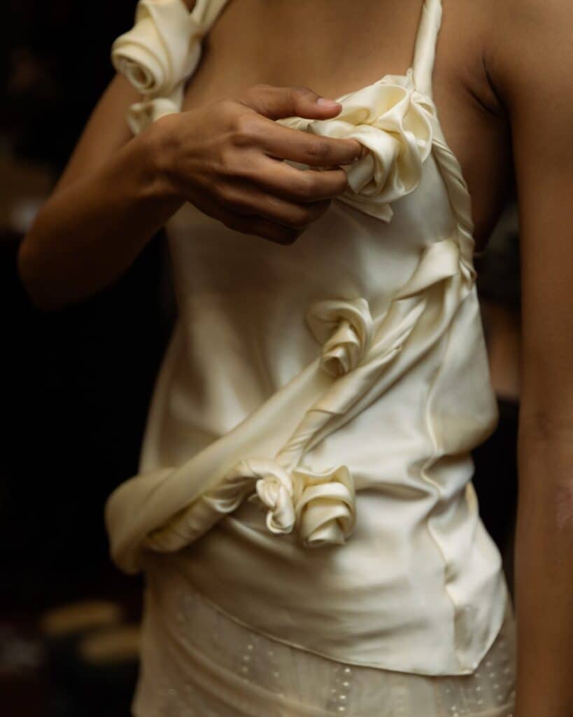 A person wearing a cream-colored satin dress adorned with fabric rose details adjusts a strap on their shoulder. The image focuses on the upper body and hand, with a dark, blurred background.