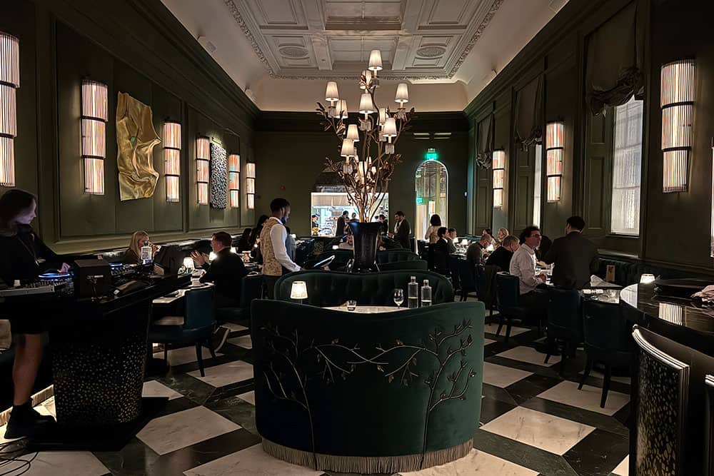 Elegant restaurant with dark green walls, checkered marble floor, and ambient lighting. Perfect for those seeking Where To Eat During London Fashion Week, it features a large decorative tree centerpiece and sophisticated wall art.