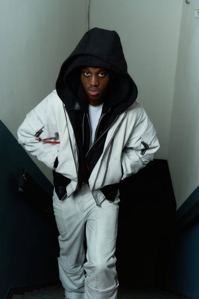 A person wearing a white jacket and pants with a black hood up stands on a staircase, leaning slightly forward with hands in pockets, looking directly at the camera. The background is a plain wall.