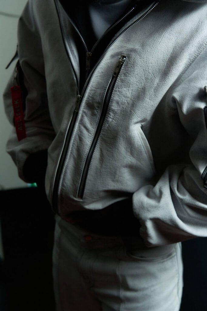 Person wearing a light gray textured jacket with multiple zippered pockets and matching pants. The jacket has a red tag on the sleeve, and the person has their hand resting on their midsection. Background is dark and blurred.