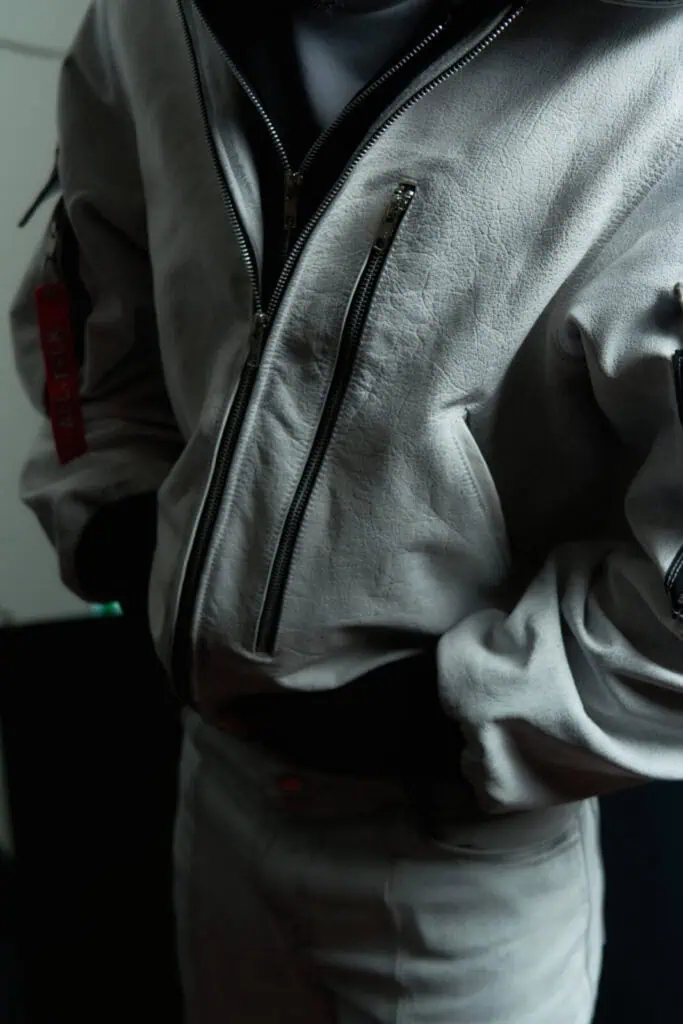 Person wearing a light gray textured jacket with multiple zippered pockets and matching pants. The jacket has a red tag on the sleeve, and the person has their hand resting on their midsection. Background is dark and blurred.