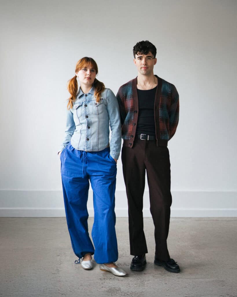 A woman in a denim jacket and loose blue pants stands next to a man wearing a patterned jacket over a black shirt and brown pants. Both face forward in a simple, light-filled room with plain walls.