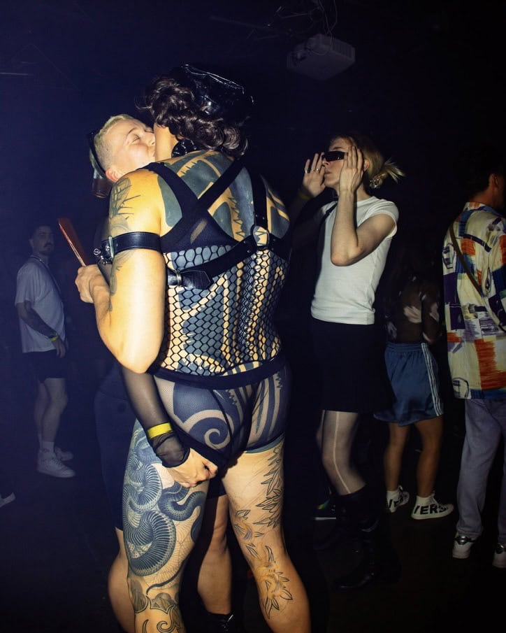 Two people kiss passionately at a club; one wears mesh lingerie and harness, showing tattoos, while the other has short hair and holds a fan. Others dance nearby, including a person in sunglasses and a white top. The scene is dimly lit.