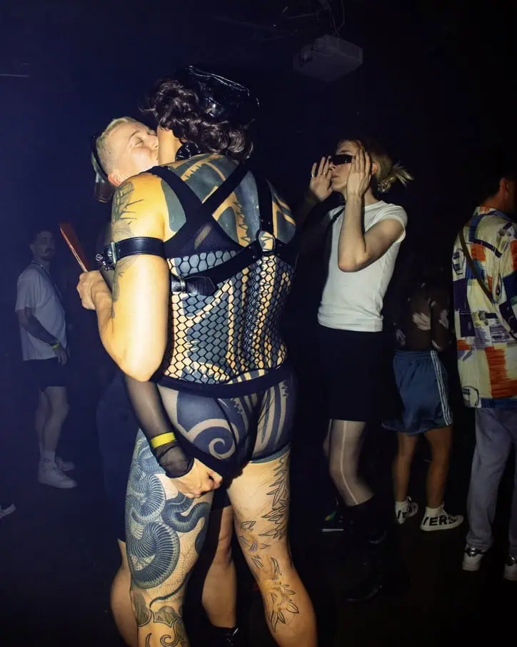 Two people kiss passionately at a club; one wears mesh lingerie and harness, showing tattoos, while the other has short hair and holds a fan. Others dance nearby, including a person in sunglasses and a white top. The scene is dimly lit.