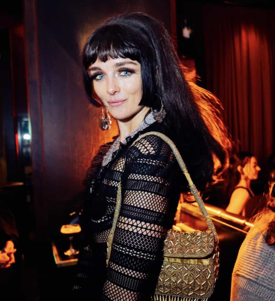 A woman with long dark hair, dramatic eye makeup, and dangling earrings wears a black mesh dress and gold woven bag, posing in a dimly lit bar or club with red curtains and people in the background.