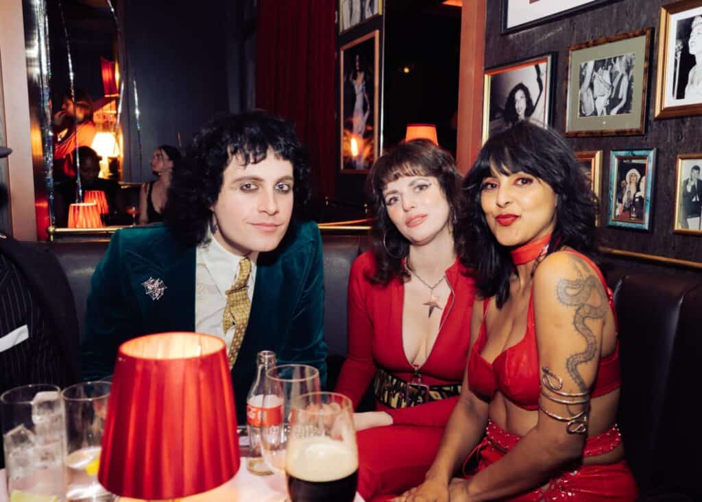 Three people in bold, retro-inspired outfits sit together in a dimly lit bar with red lamps and framed photographs on the walls, looking at the camera and smiling. Drinks are on the table in front of them.