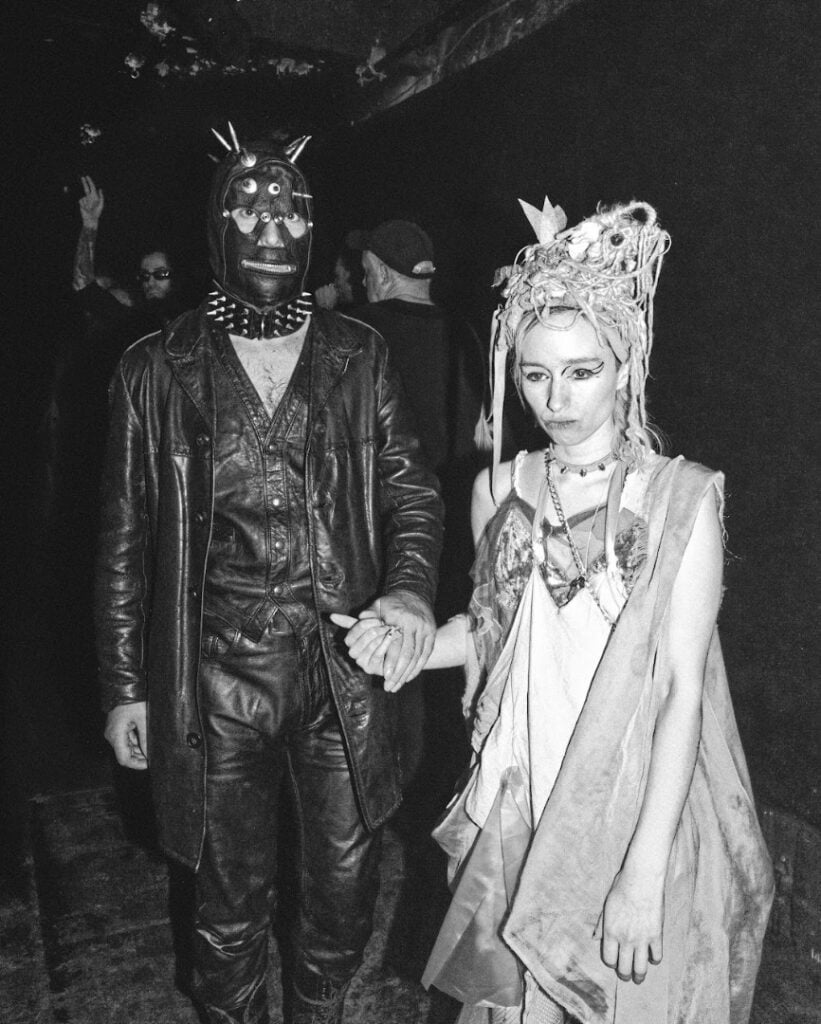 A person in a spiked leather mask and jacket holds hands with a person wearing an elaborate headpiece and layered clothing; both look serious, standing in a dimly lit environment.