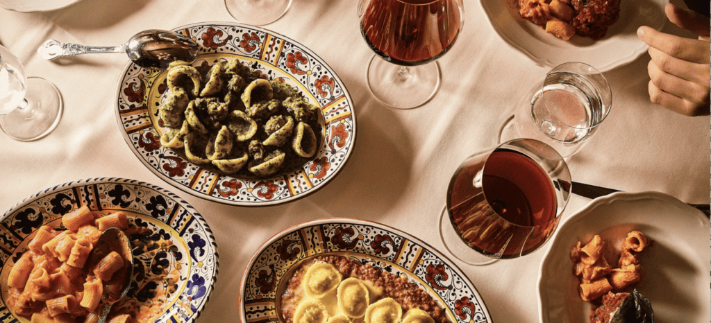 Overhead view of a table with colorful plates of pasta, glasses of red wine and water, a spoon, and a hand reaching for food—a perfect scene to inspire your list of Where To Eat During London Fashion Week.
