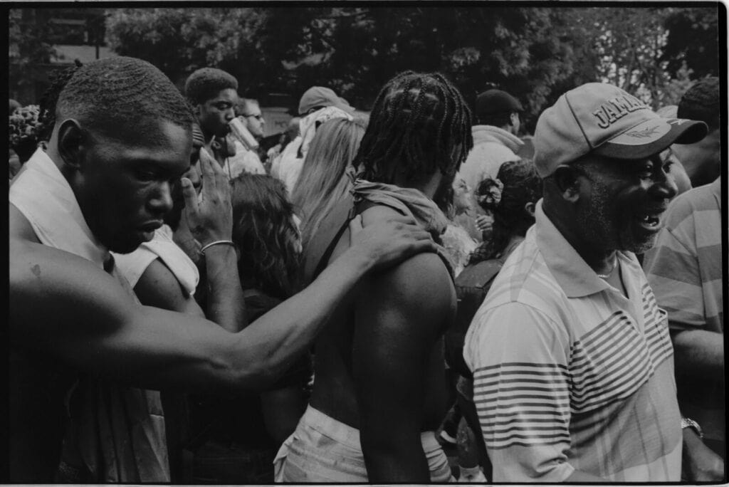 A group of people stand close together outdoors. One man rests his hand on anothers shoulder, while another man in a hat and striped shirt smiles. The scene appears lively and crowded.
