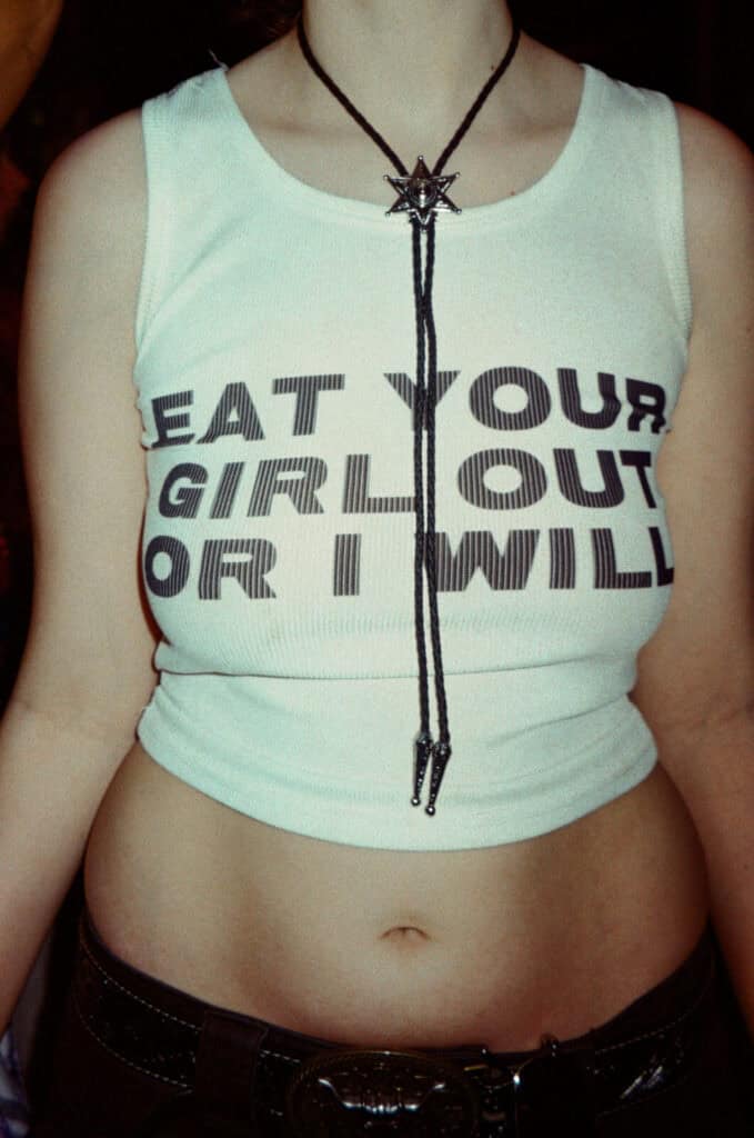 A person wearing a white sleeveless crop top with bold black text that reads, EAT YOUR GIRL OUT OR I WILL and a black cord necklace with a star-shaped pendant. Their midriff is visible.