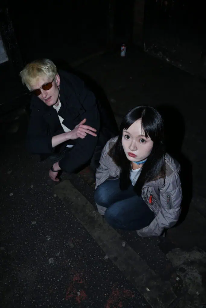 Two people crouch on a dark, worn pavement at night. One wears sunglasses and a dark coat, the other has long dark hair and a gray jacket. Both look up toward the camera, which uses a harsh flash.
