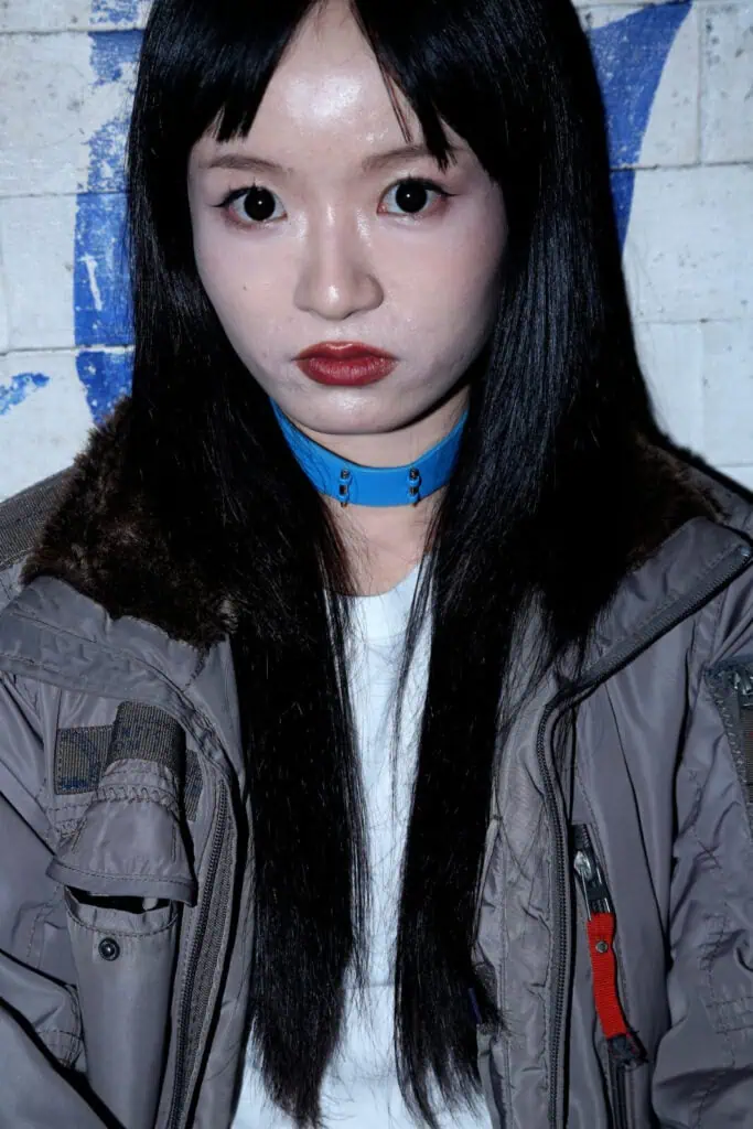 A person with long black hair and pale skin wears a gray jacket with a furry collar, a white shirt, and a blue choker. They have straight bangs and intense eye makeup, posing against a worn white wall with blue graffiti.