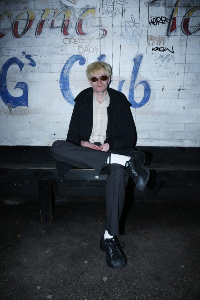 A person with blond hair, wearing sunglasses, a black jacket, and dark pants, sits on a bench against a graffiti-covered white brick wall, looking at the camera with legs crossed. The lighting is dim and moody.