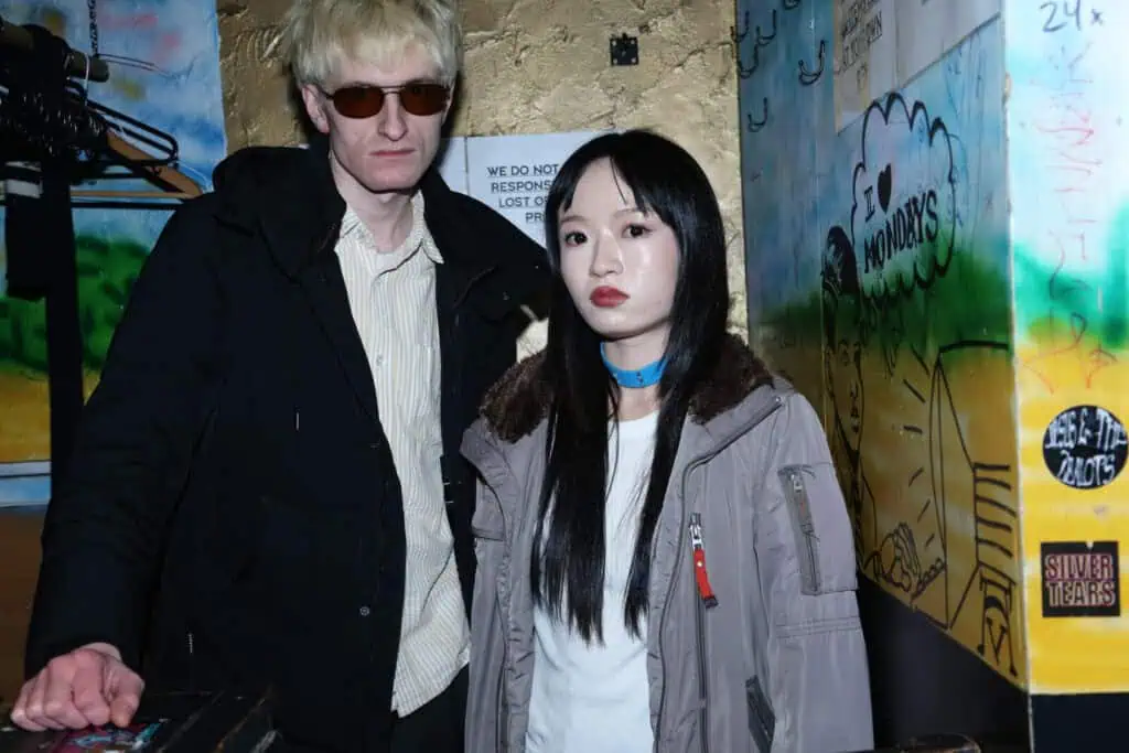 A man in sunglasses and a dark coat stands next to a woman in a gray jacket with straight black hair. They are indoors, in front of colorful graffiti and a metallic gold wall. Neither person is smiling.