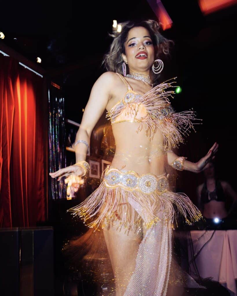 A woman in a sparkling, beaded belly dance costume performs onstage with red curtains and colorful lights in the background. She has hoop earrings, raised arms, and an expressive face.