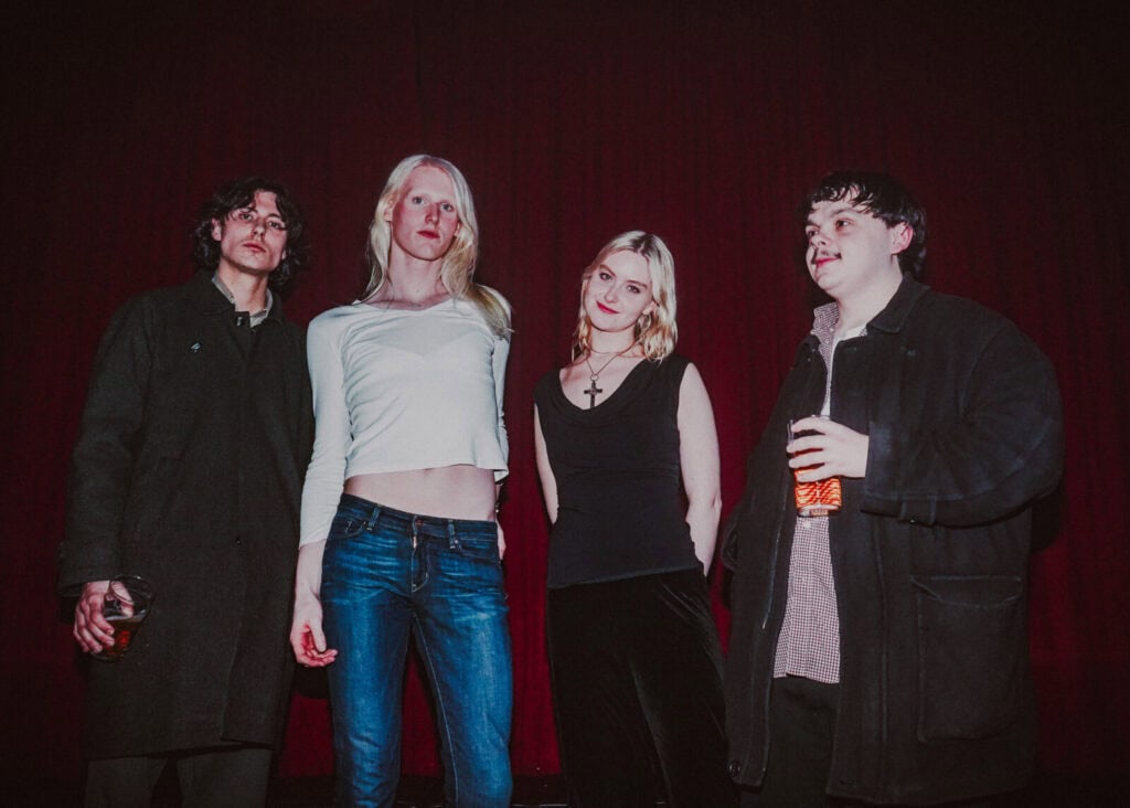 Four young adults, members of the Bathing Suits band, stand in front of a dark red curtain, casually posing for an interview. Two hold drinks, their dark, stylish clothing and the dim lighting adding to the relaxed, moody atmosphere.