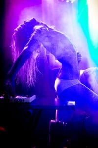 A person in underwear arches backward dramatically while operating DJ equipment, illuminated by vibrant purple and blue stage lighting and surrounded by smoke.