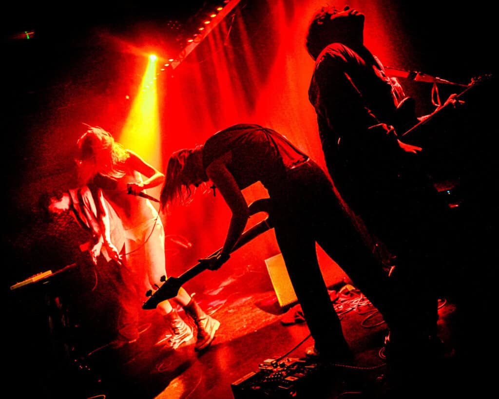 The Bathing Suits band performs energetically on stage under dramatic red lighting, with silhouettes of musicians playing guitars and a singer in motion at the microphone, capturing the excitement before their interview.