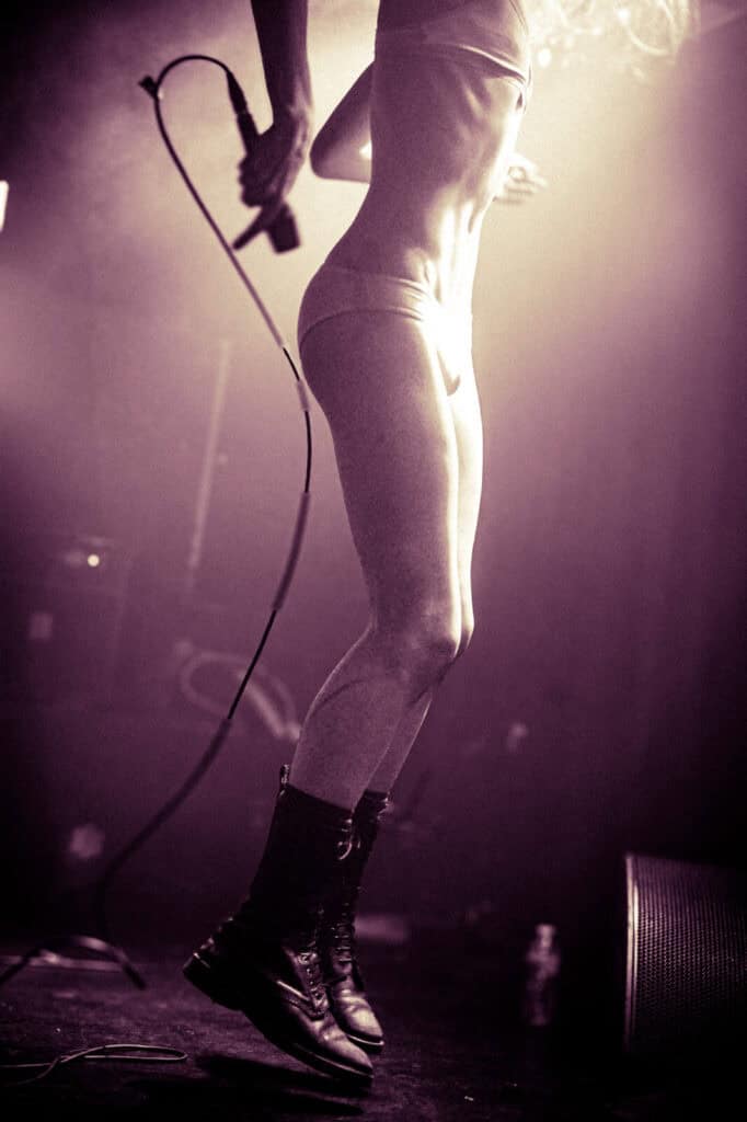 A person from the Bathings Suits band, in underwear and boots, is mid-jump on stage holding a microphone with a trailing cord. Dramatic purple, hazy lighting highlights their energetic movement—perfect for an interview moment.