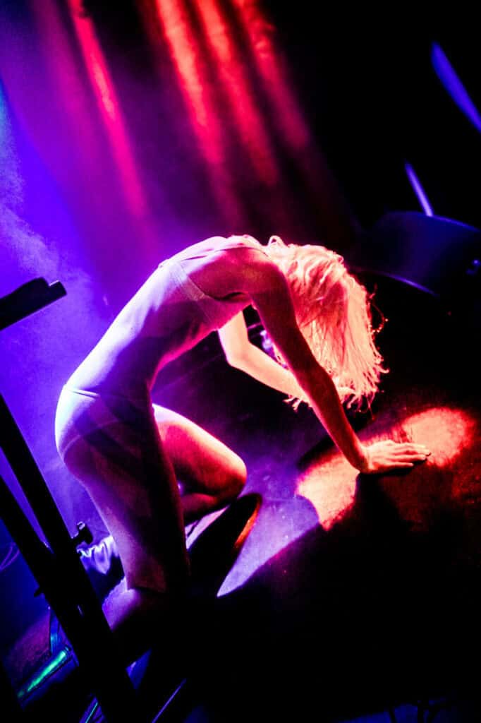 A performer with light hair in a fitted outfit kneels dramatically on stage under colorful red and purple lighting, evoking the intensity seen in a Bathings Suits band interview, with their head bowed and one hand on the floor.