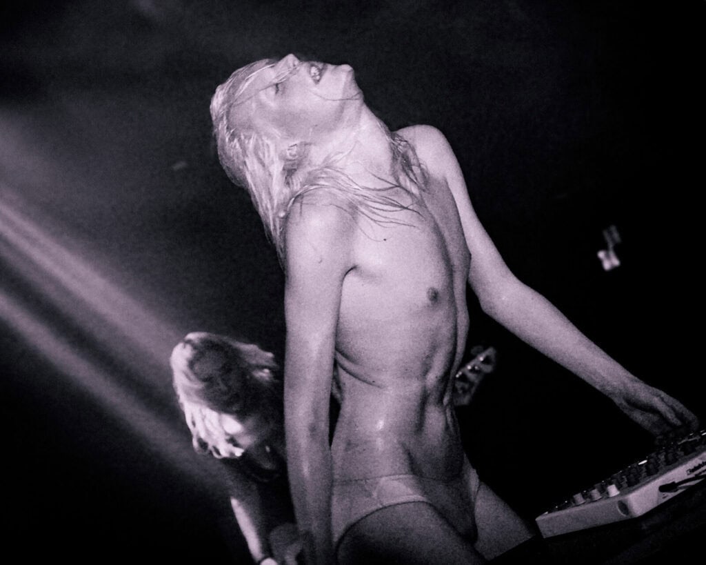 A person with long wet hair, wearing minimal clothing, leans back dramatically while performing on stage with a keyboard. Another musician from Bathings Suits band plays guitar in the dimly lit background.