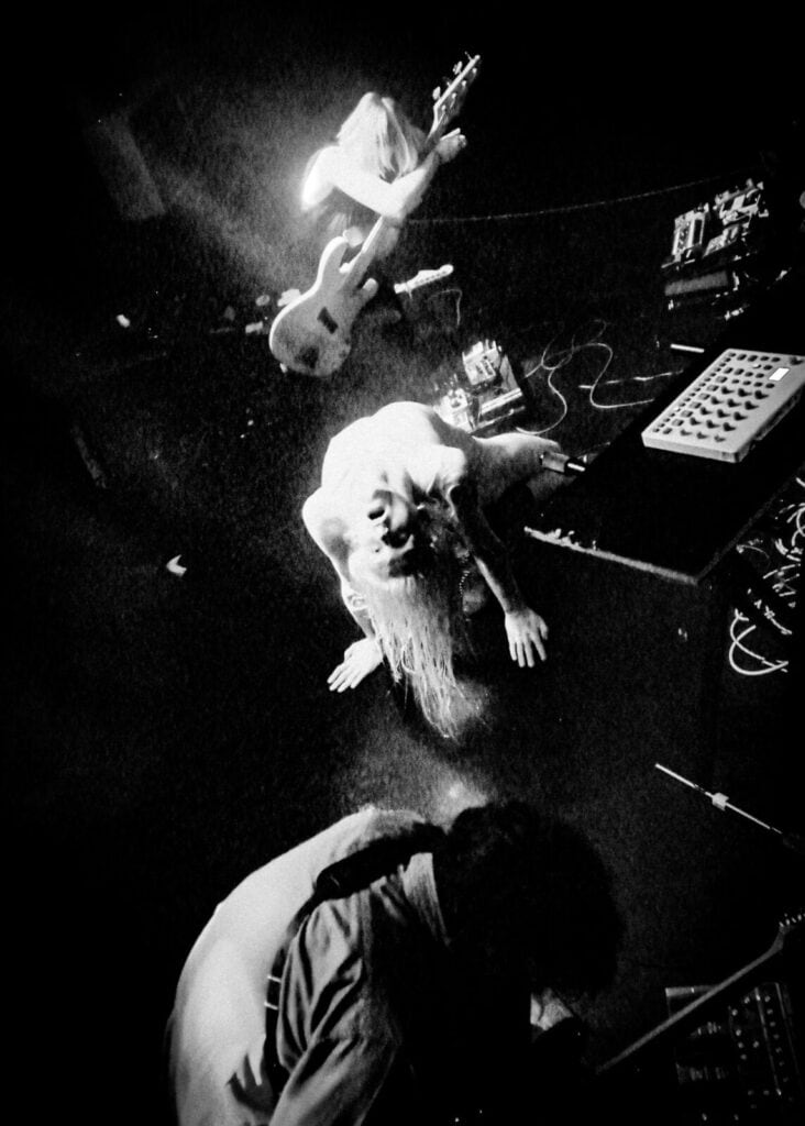 Black and white image of three Bathings Suits band members performing on stage, captured from above. One plays guitar, another kneels with hair hanging down, and the third bends forward amid cables and electronic equipment—perfect for an interview feature.