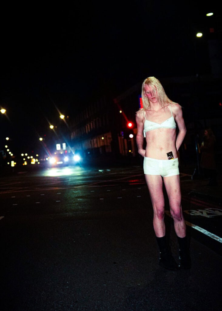 A person with long blond hair stands on a wet city street at night, wearing a white bralette, white shorts—styled like bathing suits—and black boots. Streetlights and car headlights illuminate the dark urban background, as if ready for a band interview.