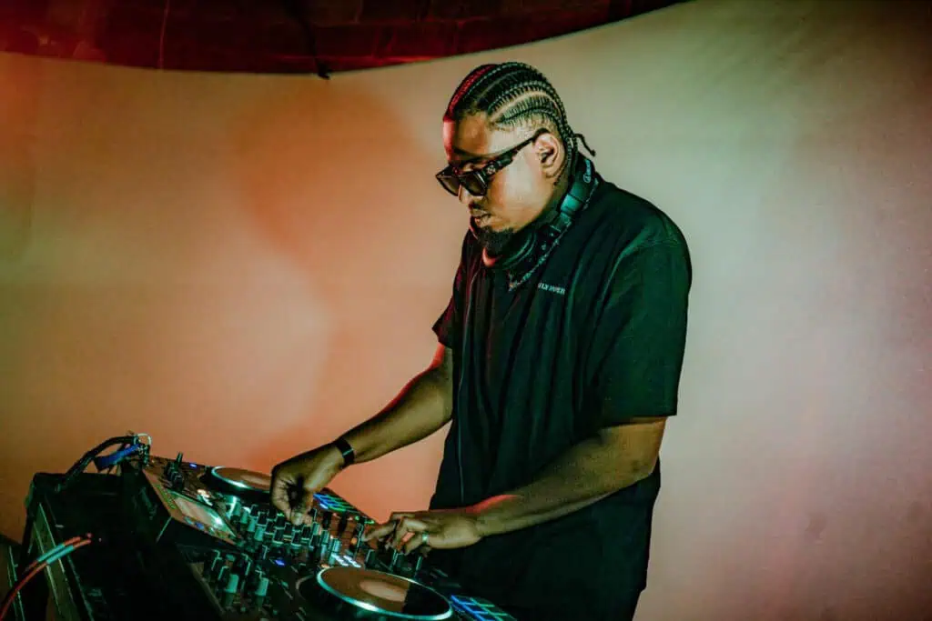 A DJ wearing sunglasses and headphones stands at a mixing console, focused on adjusting the controls. The background is softly lit in warm tones.