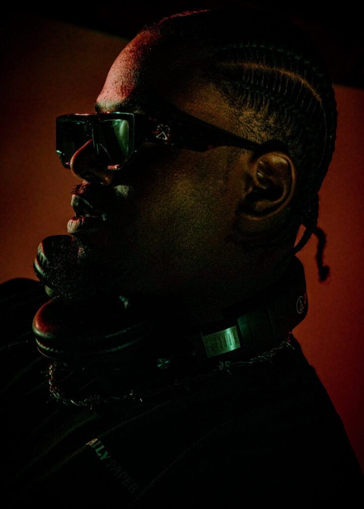 A man with braided hair and sunglasses is wearing headphones around his neck, illuminated by dramatic, low lighting with red and green tones.
