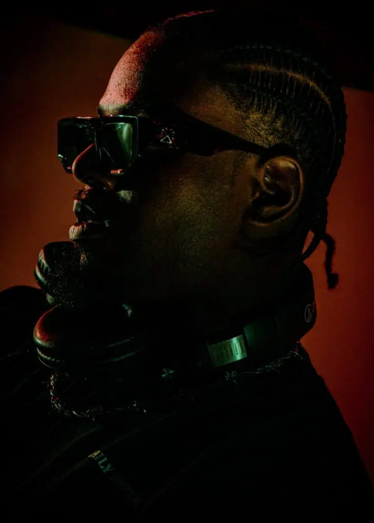A man with braided hair and sunglasses is wearing headphones around his neck, illuminated by dramatic, low lighting with red and green tones.