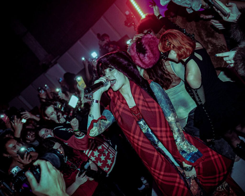 A tattooed singer wearing a red plaid coat and fur hat performs with a microphone in a crowded venue, surrounded by fans taking photos and videos with their phones. The lighting is dramatic with a red and purple glow.